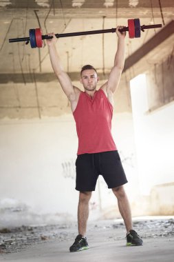 Kaslı, atletik yapılı, terk edilmiş bir binada ağırlık kaldıran genç bir sporcu.