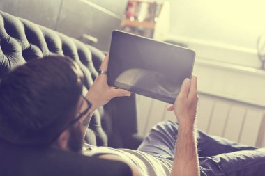 Oturma odasındaki kanepede uzanmış, tablet bilgisayarda internette sörf yapan bir erkek model.