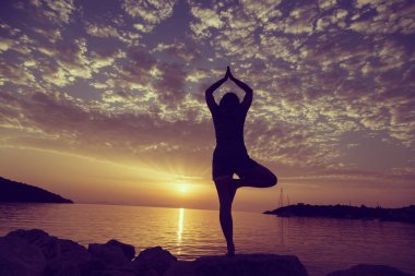 Yunanistan 'ın Syvota körfezindeki kayalık bir sahilde yoga yapan genç bir kadının silueti. Bulutlu gökyüzü, İyon denizi ve arka planda güzel bir günbatımı.