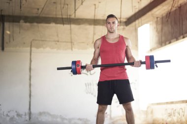 Kaslı, atletik yapılı, terk edilmiş bir binada ağırlık kaldıran genç bir sporcu.