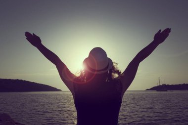 Şapka takan genç bir kadın, Syvota Bay, Yunanistan 'da sahilde duruyor, gün batımını seyrediyor ve güzellik ve özgürlüğün tadını çıkarıyor.