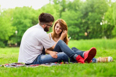 Parkta piknik battaniyesinde oturan bir çift. Adam nazikçe kızların saçlarına çiçek yerleştiriyor.