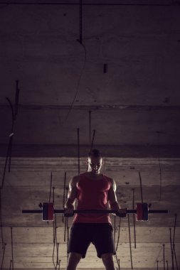 Kaslı, atletik yapılı, terk edilmiş bir binada ağırlık kaldıran genç bir sporcu.