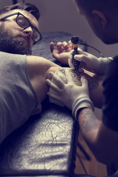 Male tattoo artist holding a tattoo gun, showing a process of making tattoos on a male tattooed model\'s arm.