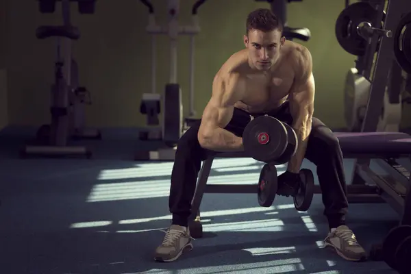 年轻肌肉建运动员在健身房里锻炼 坐在举重机升降两个哑铃 — 图库照片