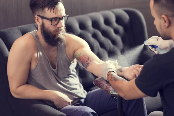 Male tattoo artist holding a tattoo gun, showing a process of making tattoos on a male tattooed model's arm.