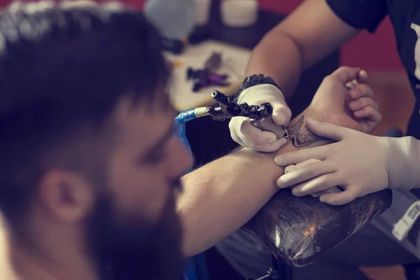 Male tattoo artist holding a tattoo gun, showing a process of making tattoos on a male tattooed model\'s arm.