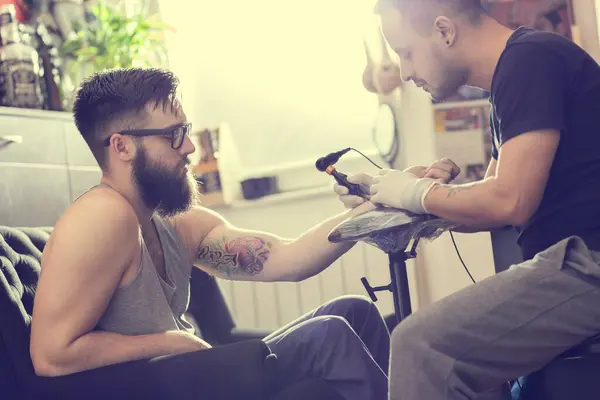 Male tattoo artist holding a tattoo gun, showing a process of making tattoos on a male tattooed model's arm.