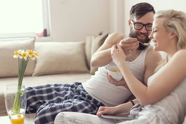 情侣们相爱地坐在客厅的沙发上 早上起床后穿着睡衣 享受着早晨的时光 男朋友用麦片喂女朋友 — 图库照片