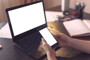 Dizüstü bilgisayar kullanan ve akıllı bir telefonu boş beyaz ekranla delip geçen kadın elleri görüntüsü. Başparmak ve telefona odaklan.