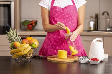 Mutfak tezgahının yanında duran önlüklü kadın meyve püresi yapmak için blender kasesini yeni kesilmiş ananasla dolduruyor.