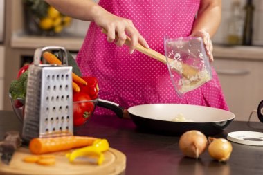 Mutfak tezgahındaki soğan kasesinin mutfak kaşığıyla doğranmış soğanı kızartma tavasına koyan kadın elleri.