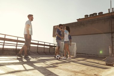 Genç arkadaşlar forma giyip, güneşli bir yaz gününde terasta futbol oynayarak eğleniyorlar. Maçtan önce ısınıyorlar.