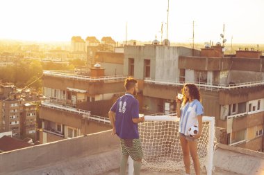 Genç çift, futbol oynadıktan sonra çatıda eğleniyor, bira içiyor ve şehirde güzel bir gün batımının tadını çıkarıyor.