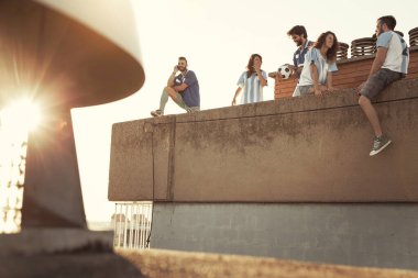 Bir grup genç arkadaş binanın çatısında futbol oynarken eğleniyor, mola veriyor ve şehrin üzerinde günbatımının tadını çıkarıyor; futbol taraftarları maçın başlamasını bekliyorlar.