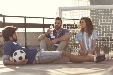 Bir grup genç arkadaş bir futbol maçından sonra terasta dinleniyor, kalenin yanında oturuyor, su içiyor ve takılıyor.