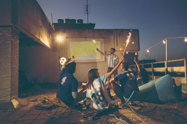 Bir grup futbol taraftarı bir binanın terasında maç izleyip gol attıktan ve maytaplarla el salladıktan sonra takımları için tezahürat yapıyorlar.