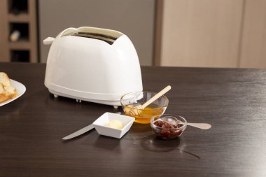 Mutfak tezgahında ekmek kızartma makinesi ve tereyağı, reçel ve bal dolu kâselerle. Tahta bal kaşığına odaklan.