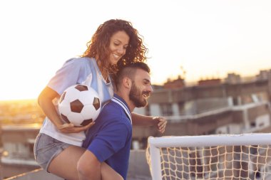 Genç çift, futbol oynadıktan sonra çatıda eğleniyor. Adam kız arkadaşını sırtında taşıyor.