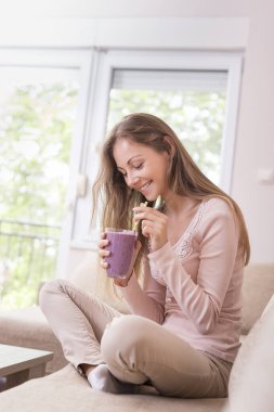 Güzel bir kız oturma odasındaki kanepede oturuyor, gülümsüyor ve elinde bir bardak ahududu ve yaban mersini kokteyli tutuyor.. 