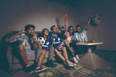 Bir grup genç arkadaş bir binanın çatısında futbol maçı izliyor, pizza yiyip bira içiyorlar, gol attıktan sonra neşeliler.