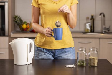 Sabahları elinde fincanla çay yapan bir kadın. Su ısıtıcısının kaynar suyuna çay ekliyor.