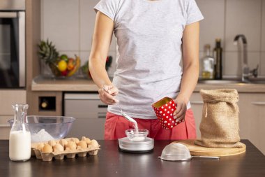 Şekeri ölçmek için mutfak terazisi kullanan kadın ellerinin ayrıntıları; pasta yapmak için malzeme ölçen kadın. Teraziye, kâseye ve kaşığa odaklan.