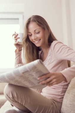 Oturma odasındaki kanepede oturan, gazete okuyan ve kahve içen genç ve güzel bir kadın.