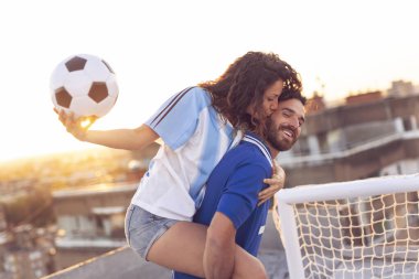 Genç çift, futbol oynadıktan sonra çatıda eğleniyor. Adam kız arkadaşını sırtında taşıyor.