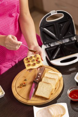 Kadın ellerinin bir sandviçi tuttuğu ve üzerine kaşık ile ketçap koyduğu yüksek açılı manzara; kahvaltıda sandviç makinesinde sıcak sandviç yapan kadın.