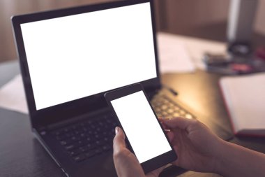 Dizüstü bilgisayar kullanan ve akıllı bir telefonu boş beyaz ekranla delip geçen kadın elleri görüntüsü. Başparmağına odaklan.