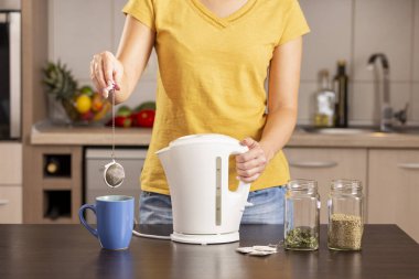 Sabahları elinde çaydanlıkla çay yapan ve çay fincanındaki kaynar suya çay poşeti ekleyen bir kadın.