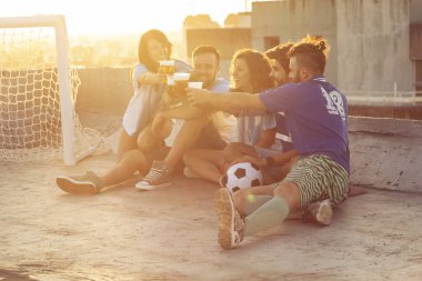 Bir grup genç bir binanın çatısında oturuyor, forma giyiyor, futbol maçından sonra dinleniyor, bira içiyor ve kadeh kaldırıyor. Sağdakilere odaklan.