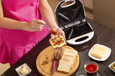 Kadın ellerinin bir sandviçi tuttuğu ve üzerine kaşık ile ketçap koyduğu yüksek açılı manzara; kahvaltıda sandviç makinesinde sıcak sandviç yapan kadın.