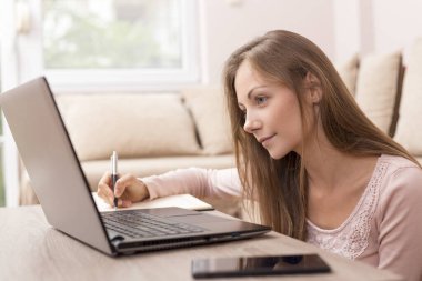 Oturma odasının zemininde oturan genç bir kadın müşterileriyle çevrimiçi bir toplantı yaparken notlar alıyor.