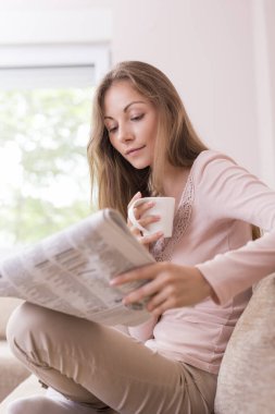 Oturma odasındaki kanepede oturan, gazete okuyan ve kahve içen genç ve güzel bir kadın.