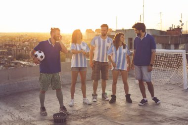 Bir futbol maçından sonra bir binanın çatısında eğlenen bir grup genç arkadaş; şehir manzarası ve arka planda kentsel günbatımıyla havada zıplıyorlar.