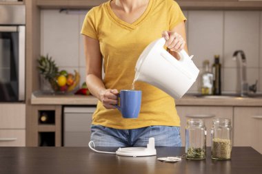 Sabahları çay yapan bir kadın, çaydanlıktan bardağa kaynar su dolduruyor.