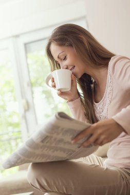 Oturma odasındaki kanepede oturan, gazete okuyan ve kahve içen genç ve güzel bir kadın.