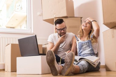 Birbirine aşık genç bir çift yeni evlerinin zemininde oturmuş dizüstü bilgisayarlarıyla internette geziniyor ve dekorasyon fikirleri için dergi okuyor.