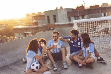 Bir grup genç bir binanın çatısında oturuyor, forma giyiyor, bir futbol maçına ara veriyor, şehirde günbatımının tadını çıkarıyor. Ortadakilere odaklan.