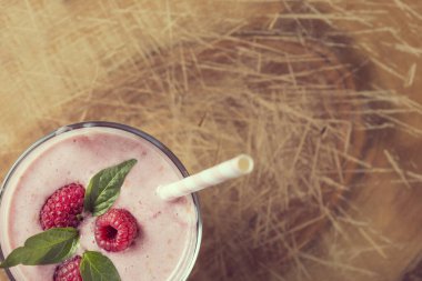 Pipetle süslenmiş ahududu smoothie bardağının üst görüntüsü, üç ahududu ve nane yaprağı, kırsal bir ahşap kesim tahtasına yerleştirilmiş.