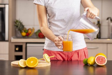 Mutfak tezgahında turunçgil sıkacağı ile hazırladıktan sonra taze sıkılmış narenciye suyunu kavanoza boşaltan kadın elleri.