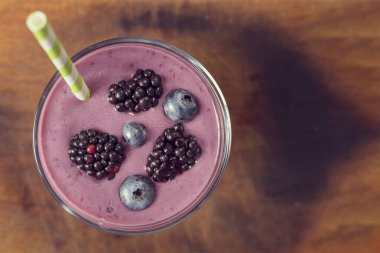 Birkaç yabanmersini ve böğürtlen ile süslenmiş, saman içen, ahşap, kırsal kesim tahtasına yerleştirilmiş bir bardak karışık meyveli milkshake 'in üst görüntüsü