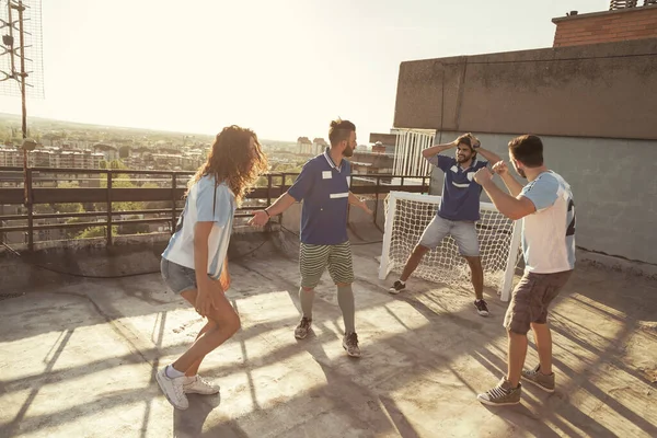 Group of young friends having fun playing football on a building rooftop terrace, winning team celebrating scoring a goal while losers mourn failure