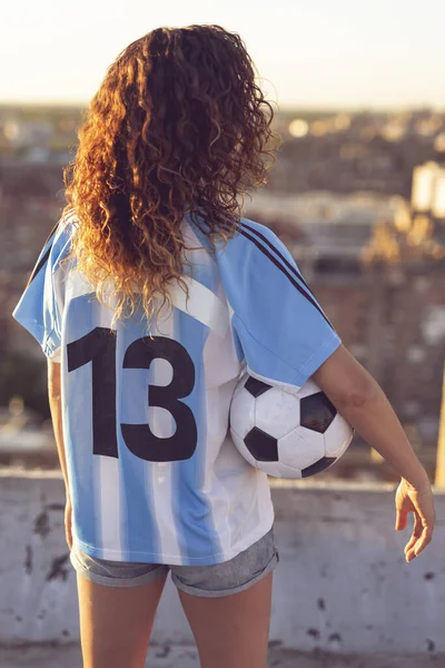 Young woman wearing a football jersey standing on a building rooftop, holding a ball and watching a sunset over the city. Focus on the jersey