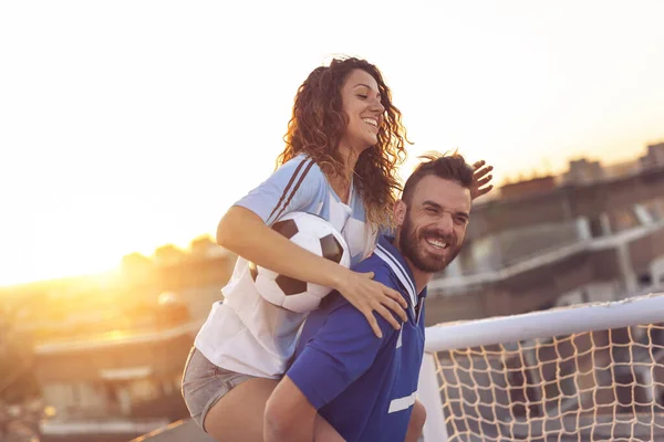 Young couple in love having fun on a building rooftop after playing football. Guy giving a piggyback ride to his girlfriend. Focus on the girlfriend