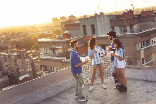 Group of young friends and football fans hanging out on a building rooftop, drinking beer before the match. Focus on the girl in the middle