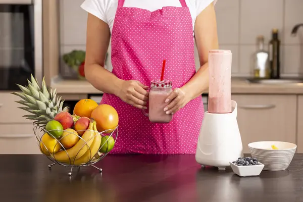 Bir kavanozda taze yapılmış ahududu smoothie tutan kadın elleri; mutfakta duran önlük giymiş kadın, kahvaltı için smoothie hazırlıyor