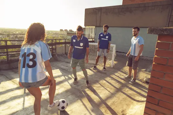 Group of young friends wearing jerseys having fun playing football on a building rooftop terrace on a sunny summer day